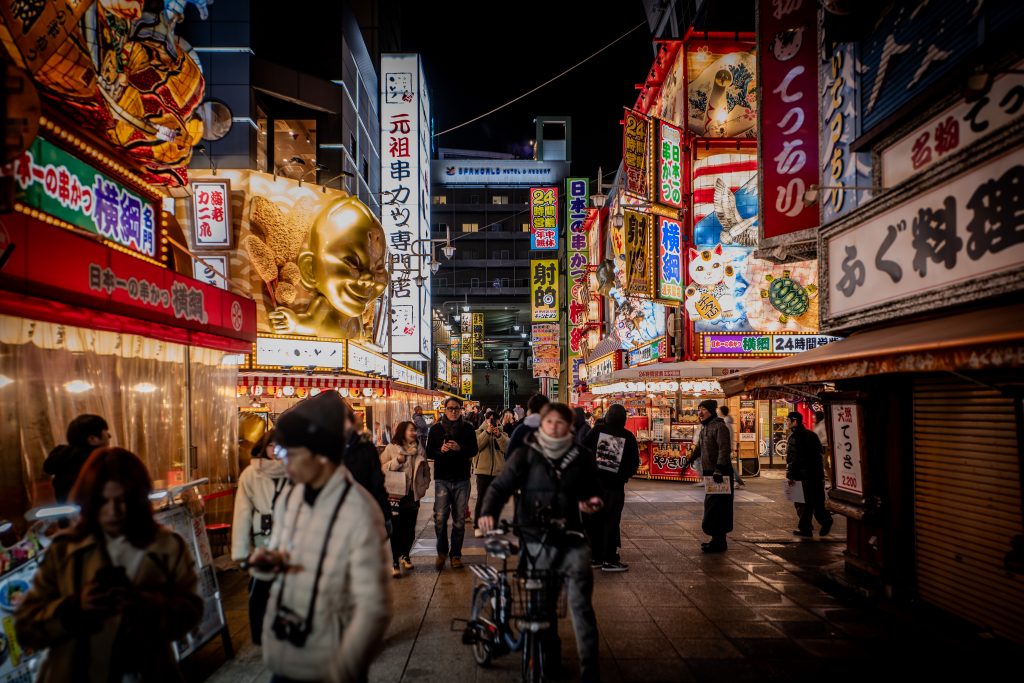 Rua movimentada em Osaka, Japão, iluminada por letreiros neon vibrantes e fachadas coloridas de restaurantes e lojas. A cena noturna mostra pedestres caminhando, turistas tirando fotos e um ciclista passando, enquanto a icônica estátua dourada de Billiken e diversos elementos tradicionais japoneses decoram o ambiente.