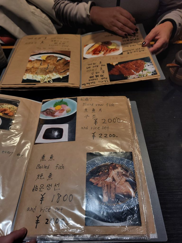 Cardápio artesanal de um restaurante japonês, feito com páginas de papel kraft plastificadas, contendo fotos dos pratos, descrições escritas à mão em diferentes idiomas e preços em ienes. As mãos de duas pessoas folheiam o cardápio, que apresenta opções como peixe cru fatiado e peixe cozido com acompanhamentos.