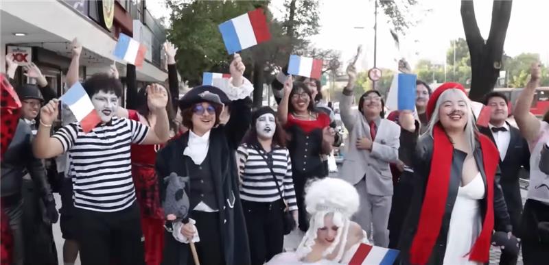 Grupo de pessoas fantasiadas com roupas e maquiagens típicas da cultura francesa, acenando bandeiras da França em uma calçada, em clima festivo