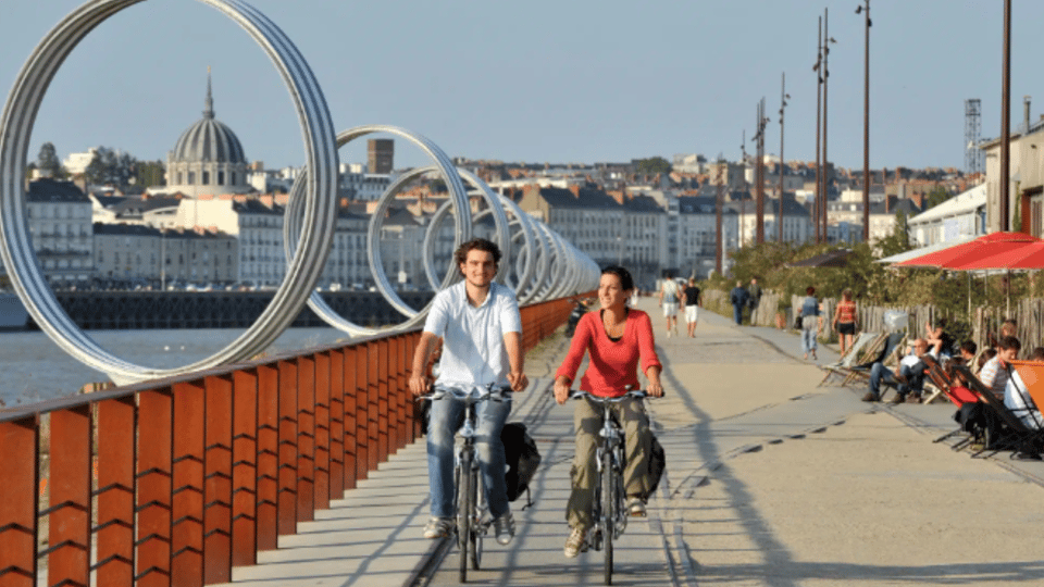 Casal andando de bicicleta ao longo do rio em Nantes, França, com esculturas circulares modernas ao fundo, representando o compromisso da cidade com sustentabilidade e espaços urbanos acessíveis