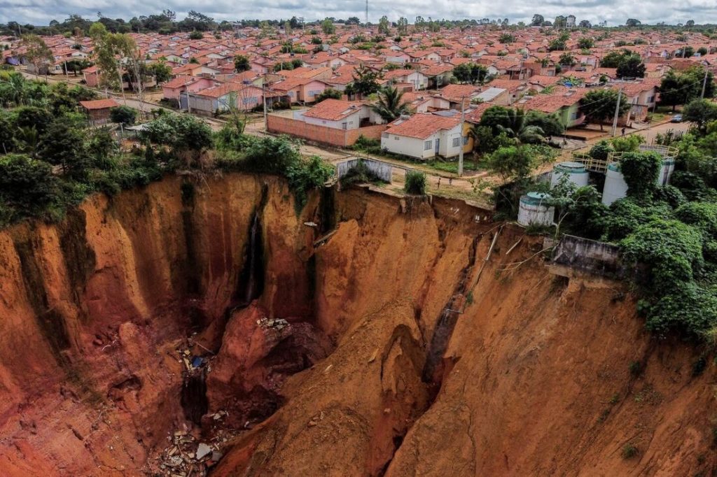 Imagem aérea mostra uma enorme cratera de erosão próxima a um bairro residencial com casas de telhado vermelho, evidenciando o risco geológico iminente para a comunidade.