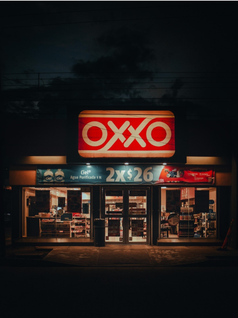 Loja de conveniência OXXO iluminada durante a noite, com destaque para o letreiro vermelho e branco e uma promoção de água mineral 2x$26 no topo da entrada.