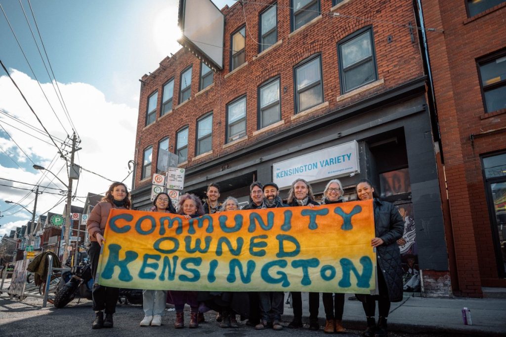  Grupo de moradores em frente a um prédio no Kensington Market, Toronto, segurando uma faixa com os dizeres 'Community Owned Kensington', representando a iniciativa comunitária de preservação local.