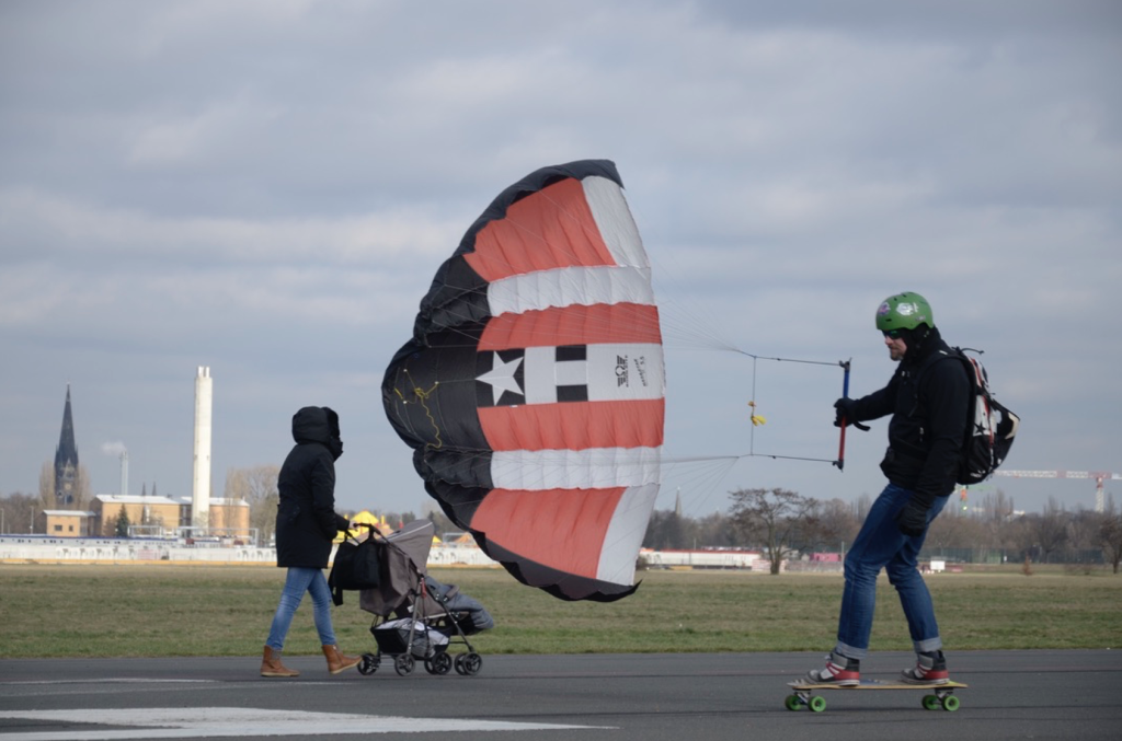 Um homem anda de longboard em uma pista de asfalto, sendo impulsionado por um pequeno paraquedas de tração com listras vermelhas, brancas e pretas. Ele usa capacete verde, mochila e roupas de frio. À esquerda da imagem, uma mulher empurra um carrinho de bebê, vestida com casaco preto e capuz. O cenário ao fundo mostra uma área aberta com grama e construções distantes sob um céu nublado.