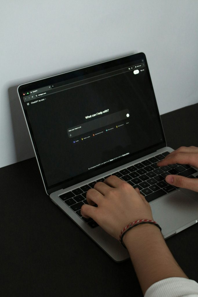 Pessoa digitando em um laptop aberto na página inicial do ChatGPT, exibindo a pergunta 'What can I help with?' em modo escuro.