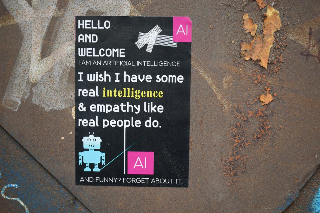 Cartaz em superfície enferrujada com mensagens irônicas sobre inteligência artificial, incluindo o texto: 'Hello and welcome, I am an artificial intelligence. I wish I have some real intelligence & empathy like real people do. And funny? Forget about it.' acompanhado de ilustrações de um robô e o logotipo 'AI' em rosa.