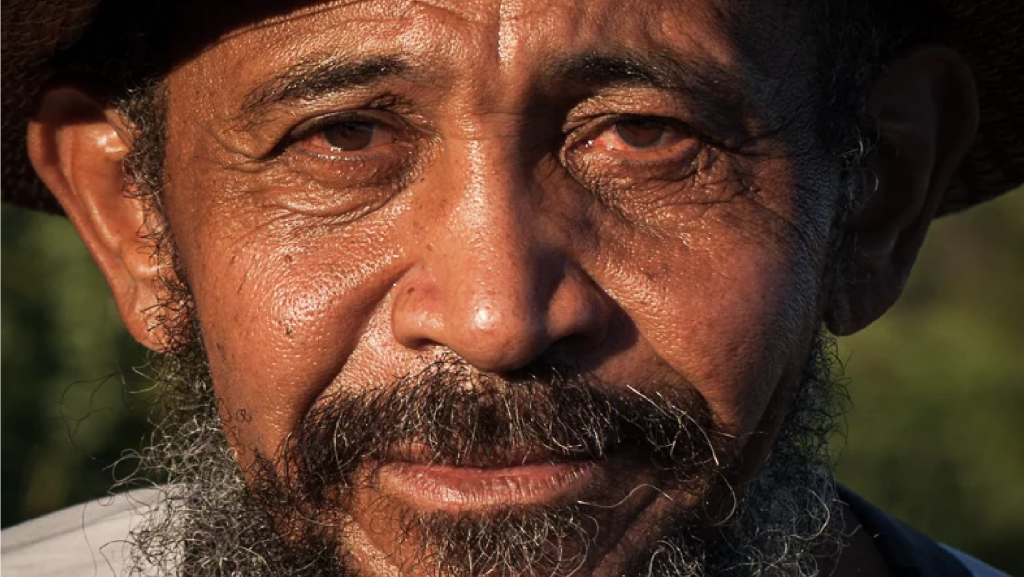 Retrato próximo de um homem mais velho com barba e chapéu, olhando diretamente para a câmera com expressão serena e profunda.